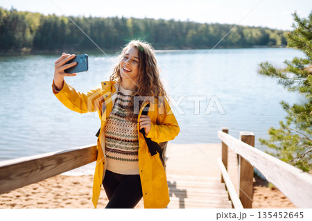 Happy woman with phone in hands taking selfie in forest. Young tourist enjoying spring day near lake 135452645