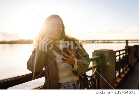 Young woman enjoys a sunny day by the river, using her smartphone while smiling in the warm sunlight Young woman enjoys a sunny day by the river, using her smartphone while smiling in the warm sunlight 135454075