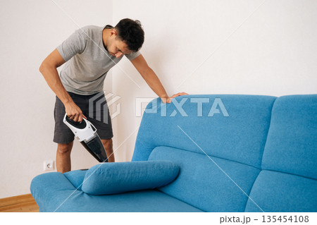 Portrait of focused young man using handheld vacuum cleaner to remove dust and dirt from blue sofa, emphasizing importance of cleanliness and hygiene in home maintenance. Portrait of focused young man using handheld vacuum cleaner to remove dust and dirt from blue sofa, emphasizing importance of cleanliness and hygiene in home maintenance. 135454108