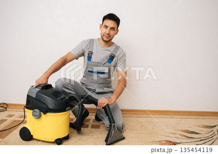 Portrait of handsome skilled cleaner male sitting on floor next to washing vacuum cleaner, holding cleaning attachment, prepared to deliver expert carpet cleaning services in home environment Portrait of handsome skilled cleaner male sitting on floor next to washing vacuum cleaner, holding cleaning attachment, prepared to deliver expert carpet cleaning services in home environment 135454119