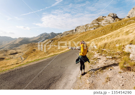 Hitchhiking woman traveler try to stop car on the mountain road. Travel and vacation theme 135454290