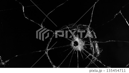 Front view photo of glass shattered by a bullet. Gunshot impact creates cracks, flying shards and dust isolated on black background. Perfect VFX overlay element or dramatic background. 135454711