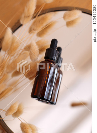 Amber glass dropper bottle reflected in a mirror with dried fluffy grass. Aesthetic skincare or essential oil concept. Amber glass dropper bottle reflected in a mirror with dried fluffy grass. Aesthetic skincare or essential oil concept. 135455089
