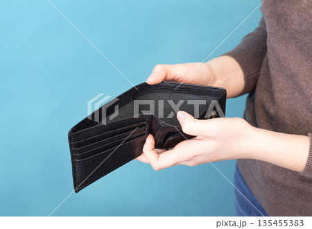 Person holding and opening an empty black leather wallet against a blue background. Person holding and opening an empty black leather wallet against a blue background. 135455383