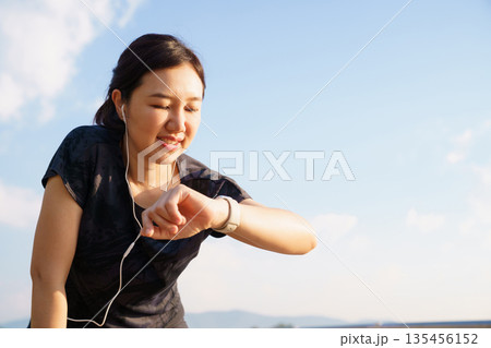 Active Asian woman checking her fitness tracker after a run outdoors. Concept of workout recovery, performance monitoring, healthy living, self care and active lifestyle. Active Asian woman checking her fitness tracker after a run outdoors. Concept of workout recovery, performance monitoring, healthy living, self care and active lifestyle. 135456152