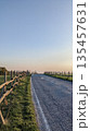 Empty rural road in Italy at golden hour, bordered by wooden fences and grassy fields, leading toward the horizon under a clear evening sky. 135457631