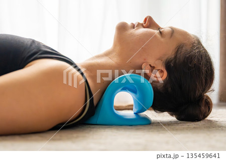 Young woman lies on the floor using a blue cervical traction device to stretch the neck. Home therapy for neck pain, muscle tension and spine decompression. 135459641