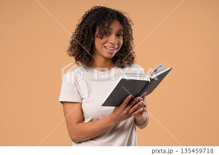 Young african woman reading book with focused smile on beige. Discovery time 135459718