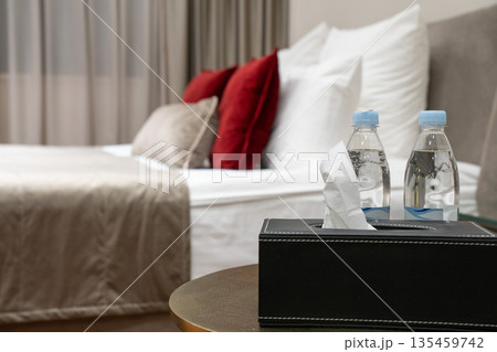 Hotel bedside table with bottled water and tissue box in a modern guest room, featuring a comfortable bed, decorative pillows and soft lighting 135459742