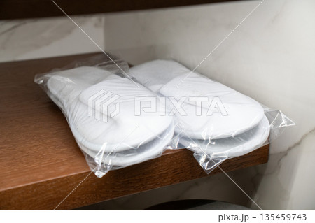 Pair of white hotel slippers individually wrapped in plastic, placed on a wooden shelf in a hotel room wardrobe 135459743