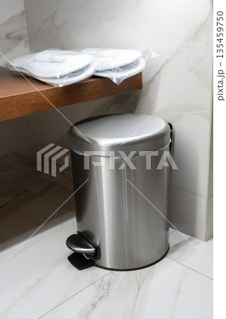 Stainless steel pedal trash can placed in a hotel bathroom under a wooden shelf with wrapped slippers 135459750