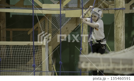 Young girl carefully climbing rope bridge in indoor adventure park. Creative Young girl carefully climbing rope bridge in indoor adventure park. Creative 135460591