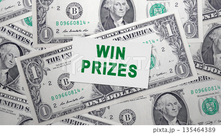 A white card with the message WIN PRIZES in green text sits atop a pile of US one-dollar bills, representing concepts of luck, winning, rewards, and financial incentives 135464389