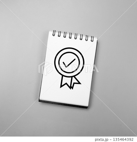 High angle shot of a white spiral notebook featuring a black line art quality seal icon with a check mark, set against a solid grey background. Perfect for business and quality control concepts High angle shot of a white spiral notebook featuring a black line art quality seal icon with a check mark, set against a solid grey background. Perfect for business and quality control concepts 135464392