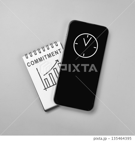Top view flat lay of a notebook with 'COMMITMENT' and a growth graph under a smartphone displaying a clock icon, representing time management and business success 135464395