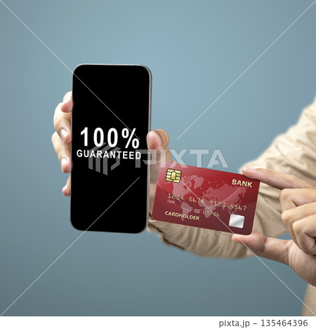 A person holds a mobile phone displaying a 100% guaranteed message next to a red credit card, representing secure digital payments, banking trust, and online shopping confidence A person holds a mobile phone displaying a 100% guaranteed message next to a red credit card, representing secure digital payments, banking trust, and online shopping confidence 135464396
