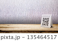 A white spiral desk calendar with 'Save The Date' text stands on a wooden table against a textured wall background, offering plenty of copy space for event announcements and reminders 135464517
