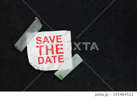 A conceptual image featuring the text 'Save the Date' in red on a crumpled piece of white paper, taped to a dark textured background with translucent tape. Great for event reminders 135464522