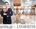 Indonesian southeast asian businessman in a formal suit holds a red circular sign with the number 53, standing in a blurred modern airport terminal or corporate office lobby 135464556