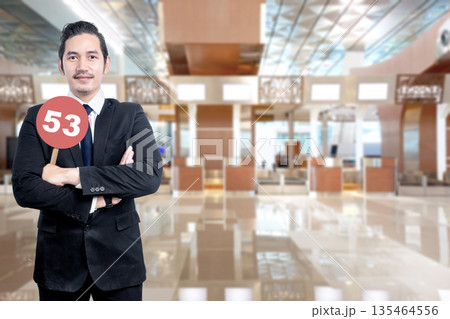 Indonesian southeast asian businessman in a formal suit holds a red circular sign with the number 53, standing in a blurred modern airport terminal or corporate office lobby 135464556