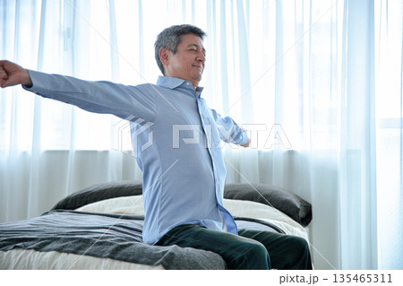 ベッドに座って伸びをするシニア男性 135465311