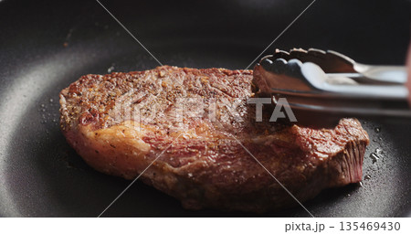 Closeup shot of man hands flip ribeye steak on non stick pan with steel tongs 135469430