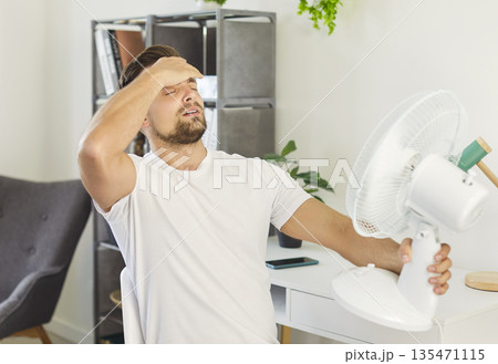 Exhausted man holding electric fan and wiping sweat from forehead while suffering from summer heat. 135471115