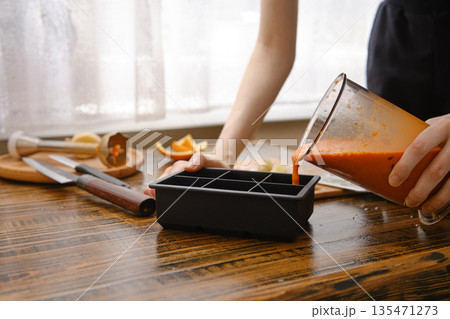 A person is pouring a blend of sea buckthorn, orange and ginger into a silicone ice tray in a kitchen in the morning A person is pouring a blend of sea buckthorn, orange and ginger into a silicone ice tray in a kitchen in the morning 135471273