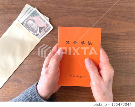 木製テーブルで年金手帳を持つ高齢女性の手と紙幣 木製テーブルで年金手帳を持つ高齢女性の手と紙幣 135473044