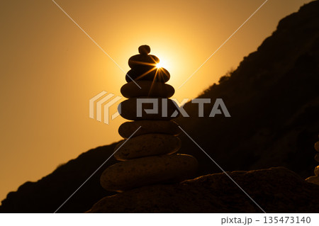 Cairn, sunset, balance. Stacked zen stones creating a silhouette with a dramatic sunburst on a rocky hillside. Cairn, sunset, balance. Stacked zen stones creating a silhouette with a dramatic sunburst on a rocky hillside. 135473140