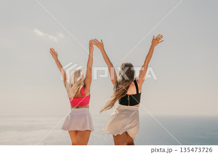 Two women are standing on a beach, one wearing a pink top and the other wearing a black top. They are both smiling and raising their arms in the air, as if they are celebrating something. Two women are standing on a beach, one wearing a pink top and the other wearing a black top. They are both smiling and raising their arms in the air, as if they are celebrating something. 135473726