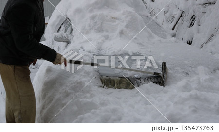 unrecognizable man uses a long-handled scraper to clear the front of a gray car, covered in deep snow after a severe storm. Clearing a car of heavy snow, starting with the front bumper and headlights unrecognizable man uses a long-handled scraper to clear the front of a gray car, covered in deep snow after a severe storm. Clearing a car of heavy snow, starting with the front bumper and headlights 135473763