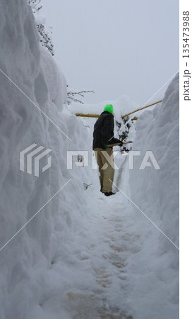 A man in dark clothing clears a path through high snowdrifts, creating a snow tunnel. The aftermath of an abnormal snowfall in a village. Manually clearing a path from heavy snow. A man in dark clothing clears a path through high snowdrifts, creating a snow tunnel. The aftermath of an abnormal snowfall in a village. Manually clearing a path from heavy snow. 135473988