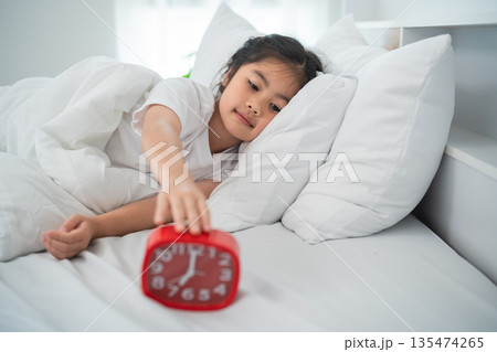 Young Girl Waking Up in Cozy Bed While Reaching for Red Alarm Clock Surrounded by Soft Pillows and Bright Morning Light in a Cheerful Bedroom Setting 135474265