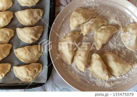 Dough dumplings arranged on a baking tray and a plate. The dumplings are uncooked and dusted with flour, ready for cooking. 135475079