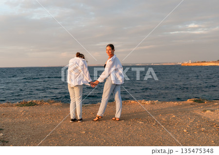 Women Beach Sunset Holding Hands - Two women standing on the beach at sunset, holding hands. Women Beach Sunset Holding Hands - Two women standing on the beach at sunset, holding hands. 135475348