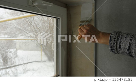 A hand with a brush begins to paint with white paint the plastered slope of a small rectangular window near a gray wall, with a view of the winter garden behind the glass A hand with a brush begins to paint with white paint the plastered slope of a small rectangular window near a gray wall, with a view of the winter garden behind the glass 135478183