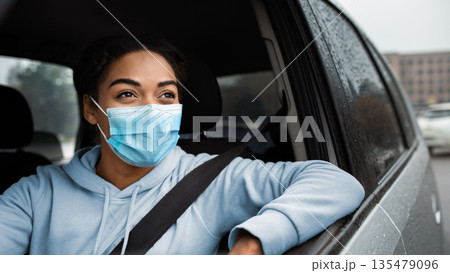 Woman holding steering wheel, driving car, insurance, driving school. Smiling millennial african american woman with seat belt, in protective mask stops and looks through open window, copy space Woman holding steering wheel, driving car, insurance, driving school. Smiling millennial african american woman with seat belt, in protective mask stops and looks through open window, copy space 135479096