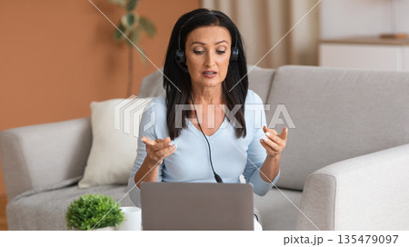 Web Conference Concept. Portrait of mature woman wearing headset sitting on couch at table, using laptop, having virtual videocall on laptop talking to webcam, explaining lesson, gesturing Web Conference Concept. Portrait of mature woman wearing headset sitting on couch at table, using laptop, having virtual videocall on laptop talking to webcam, explaining lesson, gesturing 135479097