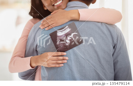 Young black pregnant woman with photo of unborn baby hugging her husband indoors, close up. Happy future mother and father with ultrasound picture of their child embracing at home Young black pregnant woman with photo of unborn baby hugging her husband indoors, close up. Happy future mother and father with ultrasound picture of their child embracing at home 135479114
