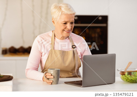 Senior Housewife Using Laptop Computer Browsing Internet For Online Recipes Cooking Dinner In Modern Kitchen At Home. Food Preparation. Retirement Hobby And Lifestyle Concept 135479121