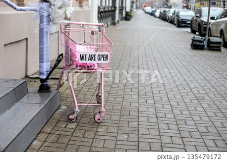 Pink Shopping Cart Displaying We Are Open Sign On Urban Sidewalk Pink Shopping Cart Displaying We Are Open Sign On Urban Sidewalk 135479172