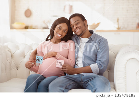 Loving black couple sitting on sofa, holding BOY and GIRL cards in hands, smiling and looking at camera at home. Young African American parents guessing their unborn baby's gender 135479198