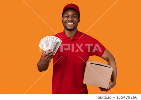 Cash On Delivery. Smiling african american male courier in red cap and uniform holding money and cardboard parcel box, carrying package, standing isolated over orange studio background 135479266