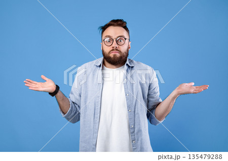 Who knows. Confused bearded man in glasses shrugging shoulders on blue studio background. Unsure millennial guy expressing uncertainty, feeling clueless, making hard choice 135479288