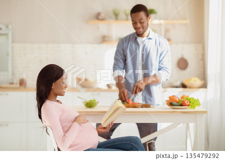 Black expectant lady reading book while her boyfriend cooking balanced breakfast in kitchen. Handsome African American guy making yummy vegetable salad for his pregnant girlfriend at home 135479292