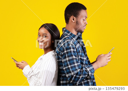 Relationships, social networks and surfing in internet. Young african american male and female stand back to back with phones, woman smiling and looking at camera, isolated on yellow background 135479293