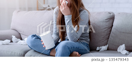 Brokenhearted young woman with bucket of ice cream crying while watching drama on TV at home. Upset lady eating for comfort because of breakup, relationship problems or stress 135479343
