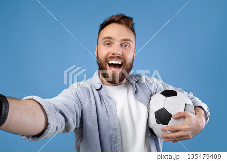 Happy soccer fan with ball taking selfie on mobile device over blue studio background. Positive football rooter making photo of himself after game, cheering for his favorite team Happy soccer fan with ball taking selfie on mobile device over blue studio background. Positive football rooter making photo of himself after game, cheering for his favorite team 135479409