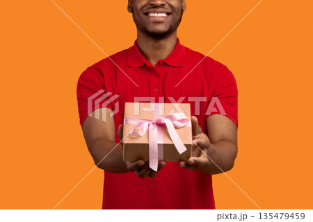 This Present Is For You. Closeup cropped view of unrecognizable smiling black bearded man holding wrapped gift box with pink ribbon, giving and showing to camera, isolated on orange studio background This Present Is For You. Closeup cropped view of unrecognizable smiling black bearded man holding wrapped gift box with pink ribbon, giving and showing to camera, isolated on orange studio background 135479459